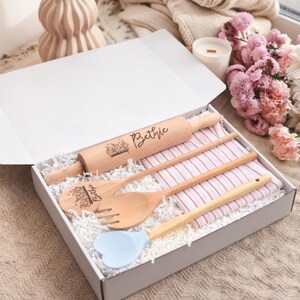 May include: A gift box containing a wooden rolling pin, two wooden spoons, a fork, and a light blue spatula. The rolling pin and one spoon are engraved with the name "Bethie" and a floral design. A red and white striped kitchen towel is also included.
