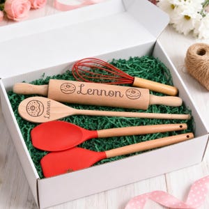 Könnte beinhalten: Eine weiße Geschenkbox gefüllt mit grünem Papier, die ein Nudelholz, einen Schneebesen, einen Löffel und zwei rote Silikonspatel enthält. Nudelholz und Löffel sind mit einem Donut und dem Namen "Lennon" graviert.