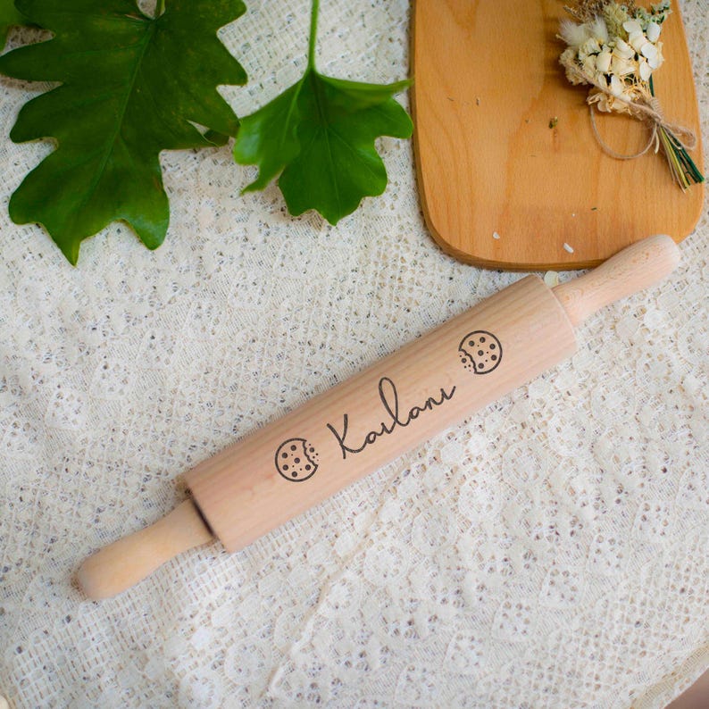 May include: A light-colored wooden rolling pin with the name "Kailani" and cookie illustrations engraved on it. The rolling pin has wooden handles and is set on a white lace surface. A wooden cutting board and greenery are in the background.