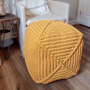 May include: A mustard yellow crocheted ottoman with a square shape and a textured surface. The ottoman is sitting on a wooden floor.
