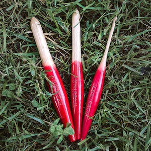 May include: Three red and white wooden crochet hooks with the text "Annebleu" on each hook. The hooks are lying on a bed of green grass.