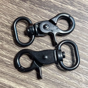 May include: Two black metal swivel clasps with a D-ring on each. The clasps are closed and ready to be attached to a strap or leash.