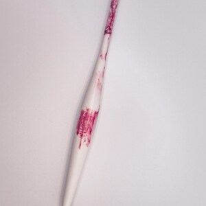May include: A white crochet hook with pink marbling. The hook is pointed at one end and has a smooth, rounded handle.