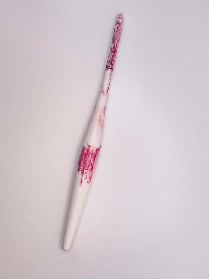 May include: A white crochet hook with pink marbling. The hook is pointed at one end and has a smooth, rounded handle.