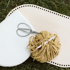 May include: A wooden oval loom with a white fabric, a ball of yellow yarn, a pair of scissors, and a crochet hook.