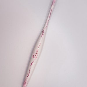 May include: A white crochet hook with pink marbling. The hook is long and slender with a pointed tip.