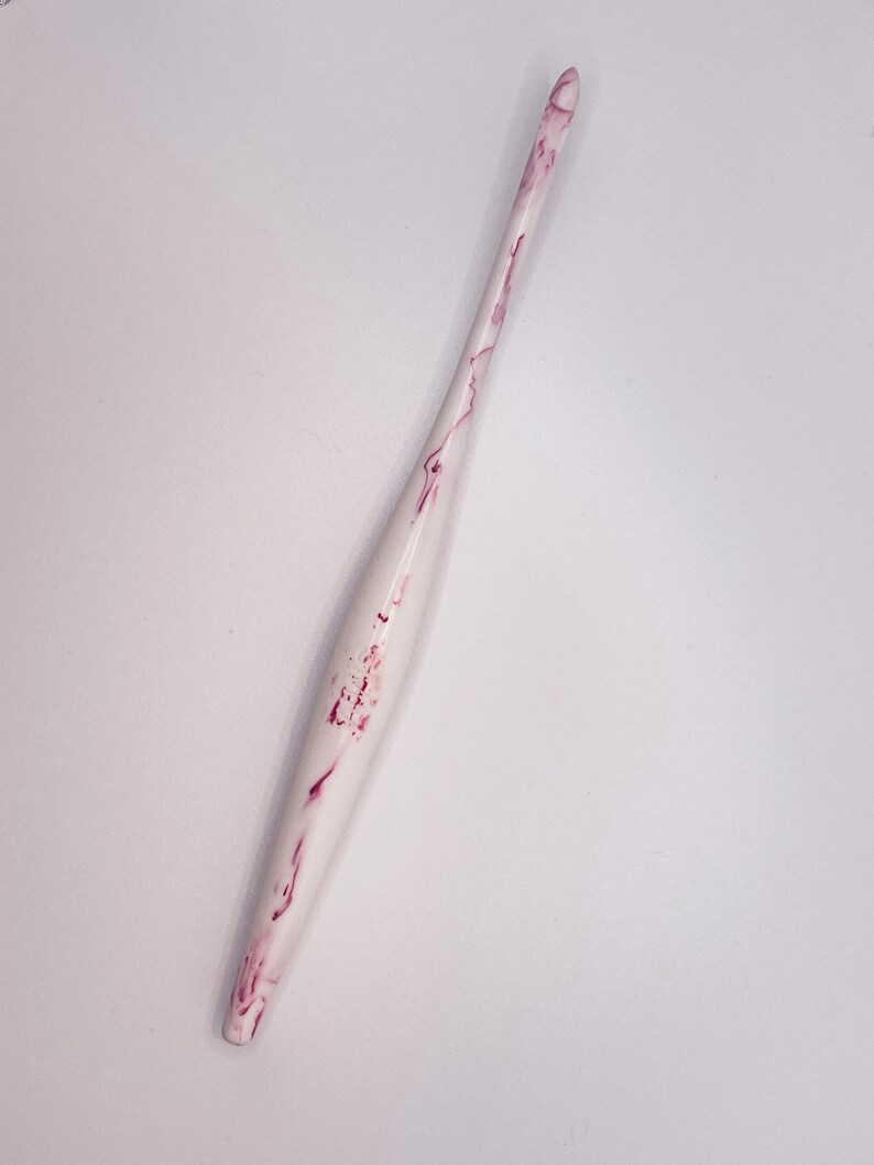 May include: A white crochet hook with pink marbling. The hook is long and slender with a pointed tip.