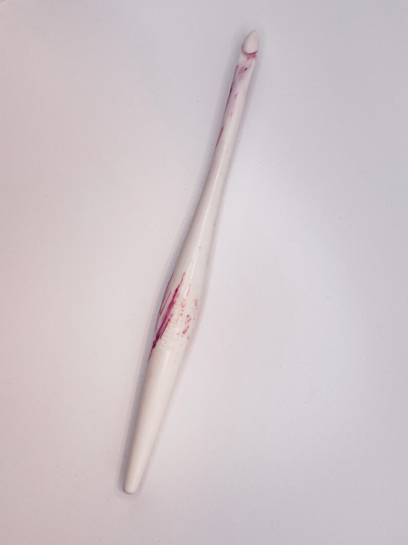 May include: White crochet hook with pink marbling. The hook has a pointed tip and a smooth handle.