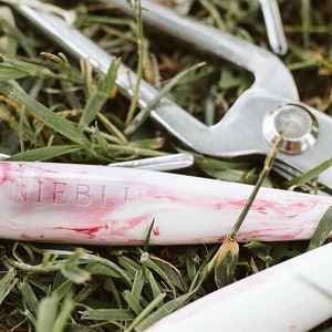 May include: A close-up of a white and pink marbled pen with the text "Annie Blue" engraved on the side. The pen is lying on a bed of green grass.