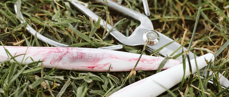 May include: A close-up of a white and pink marbled pen with the text "Annie Blue" engraved on the side. The pen is lying on a bed of green grass.
