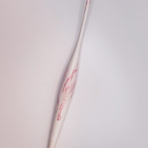 May include: A white crochet hook with pink marbling. The hook is made of plastic and has a pointed tip.