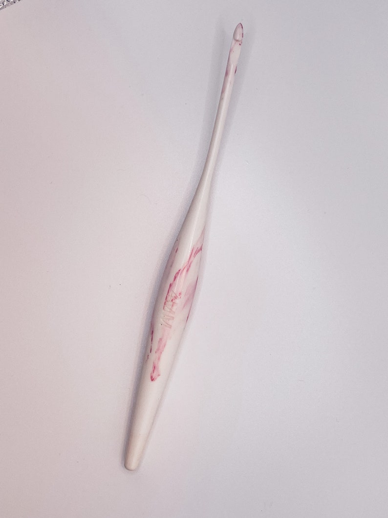 May include: A white crochet hook with pink marbling. The hook is made of plastic and has a pointed tip.