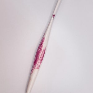 May include: A white crochet hook with a pink floral design. The hook is made of a smooth, polished material and has a pointed tip.