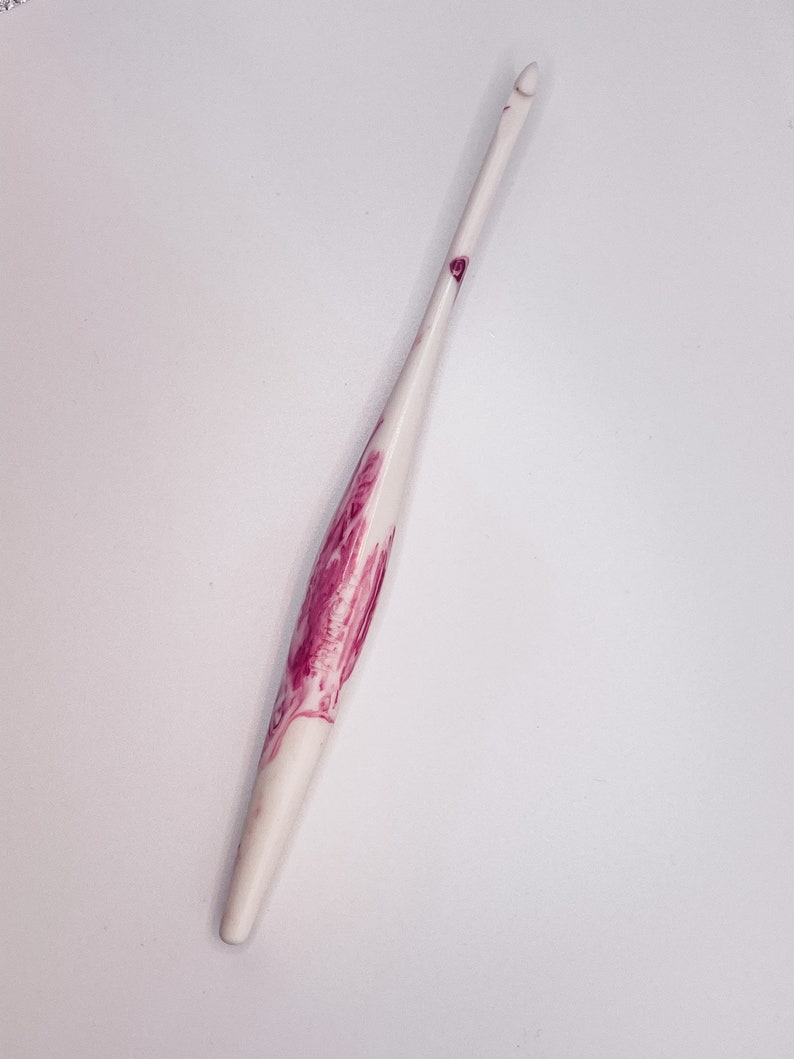 May include: A white crochet hook with a pink floral design. The hook is made of a smooth, polished material and has a pointed tip.