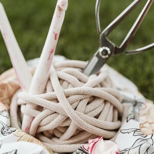 May include: A close-up of a ball of beige yarn, a pair of silver scissors, and a pink and white crochet hook. The yarn is sitting on a white fabric with a floral pattern.