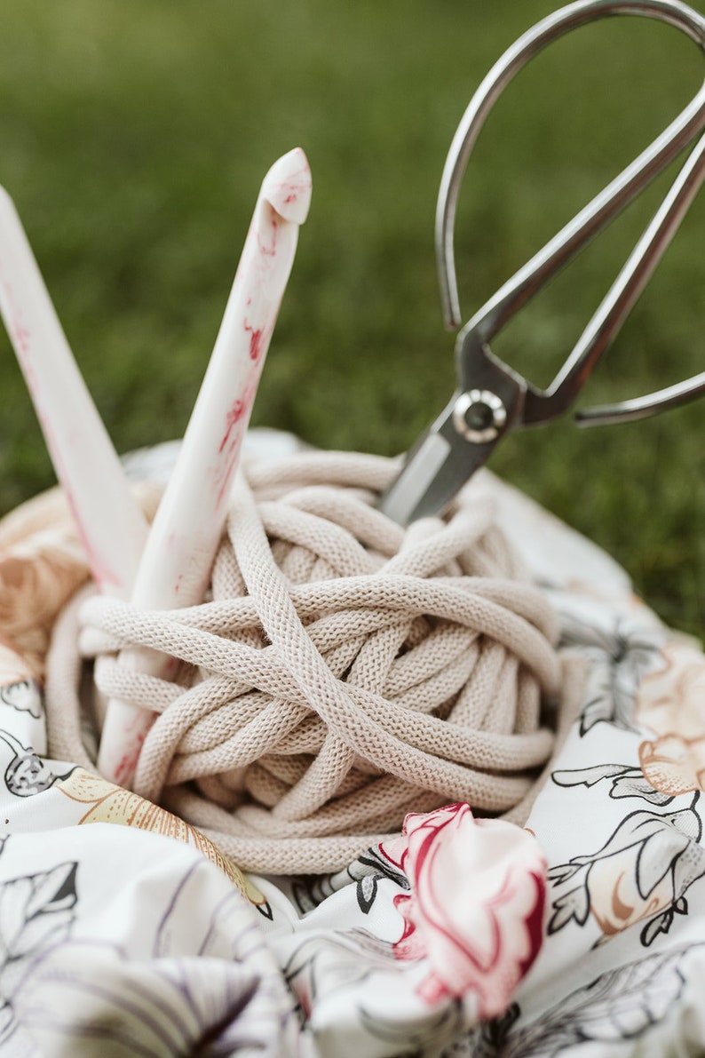 May include: A close-up of a ball of beige yarn, a pair of silver scissors, and a pink and white crochet hook. The yarn is sitting on a white fabric with a floral pattern.