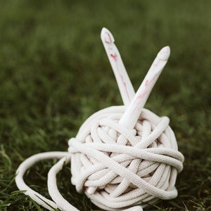 May include: A ball of beige yarn with two white crochet hooks sticking out of it. The yarn and hooks are sitting on a bed of green grass.