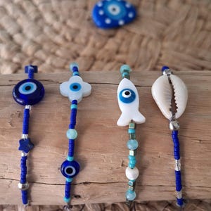 Peut inclure: Quatre breloques bleues perlées avec différents motifs : un œil protecteur, un trèfle à quatre feuilles, un poisson et un coquillage. Chaque breloque est enfilée sur un cordon de perles bleues avec des accents argentés.