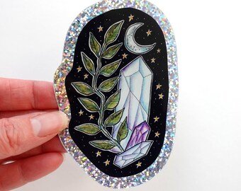 Puede incluir: Una pegatina con fondo negro que presenta un cristal, una luna creciente, estrellas y hojas verdes. El cristal es blanco y morado. La pegatina tiene un borde plateado brillante y está sujeta por una mano.