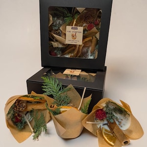 May include: A black cardboard box with a clear window containing fire-starters, with three additional fire-starters wrapped in brown paper in front. Each contains dried orange slices, cinnamon sticks, pinecones, and greenery with a label reading FIRE-STARTERS THE FIRE PIT & THE FIRE.