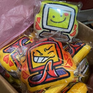 Może przedstawiać: Kolekcja pluszowych zabawek w kształcie monitorów komputerowych. Monitory są żółte z czarnymi obrysami i przedstawiają twarze z kreskówek z sercami i płomieniami. Są zapakowane w przezroczysty plastik i ułożone w kartonowym pudełku.