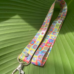 Könnte beinhalten: Ein farbenfrohes Schlüsselband mit einem sich wiederholenden Muster aus Cartoon-Figuren in Quadraten. Das Schlüsselband hat einen silberfarbenen Metallverschluss. Der Hintergrund ist ein großes grünes Blatt.