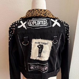 May include: A black denim jacket featuring a leopard print collar, silver metal studs on the shoulders, and various patches. The back displays patches with the text THE EXPLOITED, SORE THROAT, and DOOM, along with two metal D-rings attached to the back.