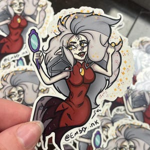 May include: A sticker featuring a cartoon illustration of a character with gray hair, wearing a red dress, and holding a mirror. The sticker has a white border and the text "@Embry.ink".