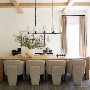 Puede incluir: Una gran mesa de comedor de madera con ocho sillas tapizadas en gris. La mesa est&aacute; puesta con un candelero negro y un jarr&oacute;n con vegetaci&oacute;n. La habitaci&oacute;n tiene paredes blancas, una ventana grande con cortinas beige y un techo de vigas de madera.