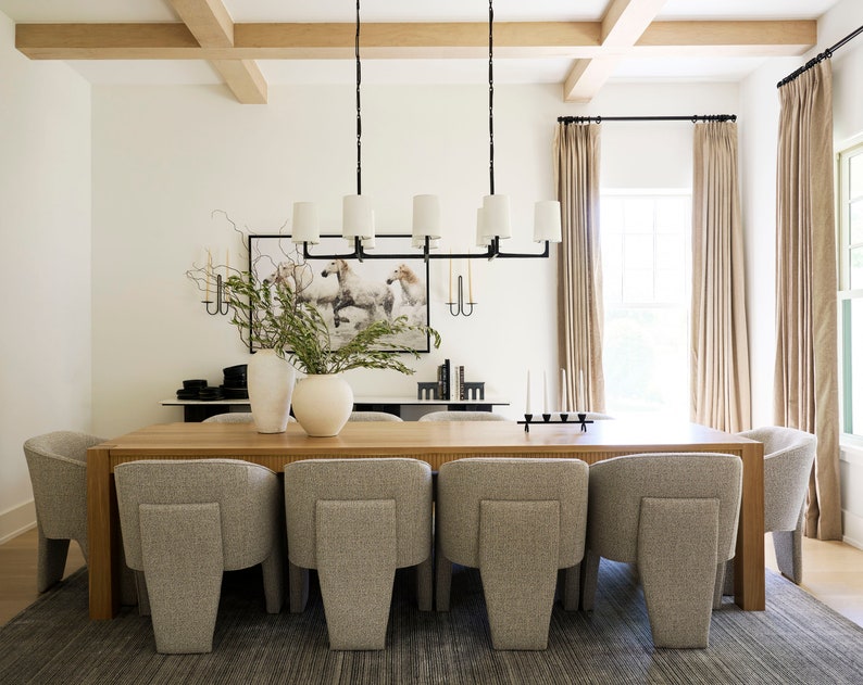 Puede incluir: Una gran mesa de comedor de madera con ocho sillas tapizadas en gris. La mesa est&aacute; puesta con un candelero negro y un jarr&oacute;n con vegetaci&oacute;n. La habitaci&oacute;n tiene paredes blancas, una ventana grande con cortinas beige y un techo de vigas de madera.