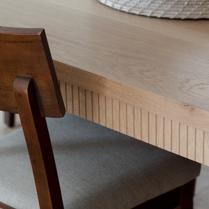 Puede incluir: Primer plano de una mesa de comedor de madera marr&oacute;n claro con un borde acanalado detallado. La mesa est&aacute; hecha de madera maciza y tiene un acabado natural. Una silla de madera marr&oacute;n con un asiento de tela gris claro es parcialmente visible en primer plano.