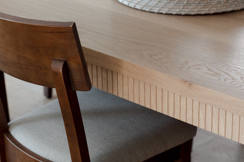 Puede incluir: Primer plano de una mesa de comedor de madera marr&oacute;n claro con un borde acanalado detallado. La mesa est&aacute; hecha de madera maciza y tiene un acabado natural. Una silla de madera marr&oacute;n con un asiento de tela gris claro es parcialmente visible en primer plano.