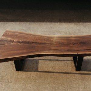 May include: A large, live edge coffee table made of dark walnut wood with a natural, wavy edge. The table is supported by two black metal legs.