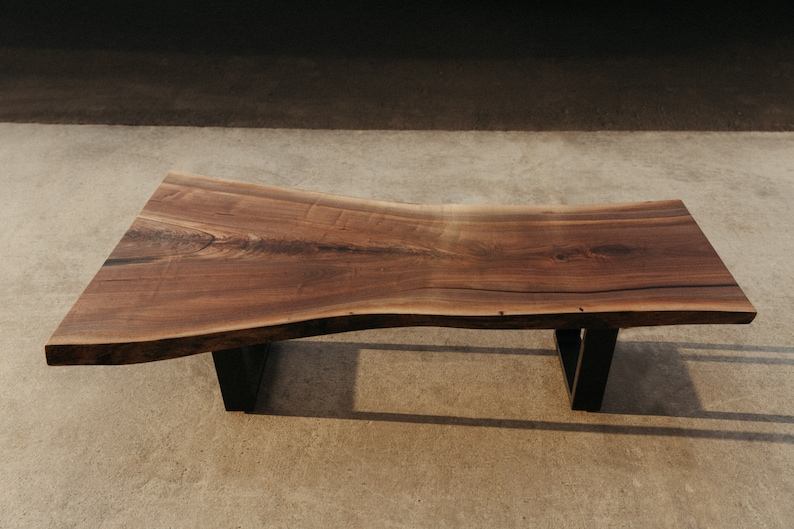 May include: A large, live edge coffee table made of dark walnut wood with a natural, wavy edge. The table is supported by two black metal legs.