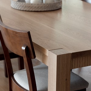 Puede incluir: Una mesa de comedor de madera con un acabado en madera clara y una bandeja tejida con jarrones de cer&aacute;mica blanca en la parte superior. La mesa tiene una forma rectangular y est&aacute; rodeada por cuatro sillas con marcos de madera marr&oacute;n y asientos de tela gris claro.