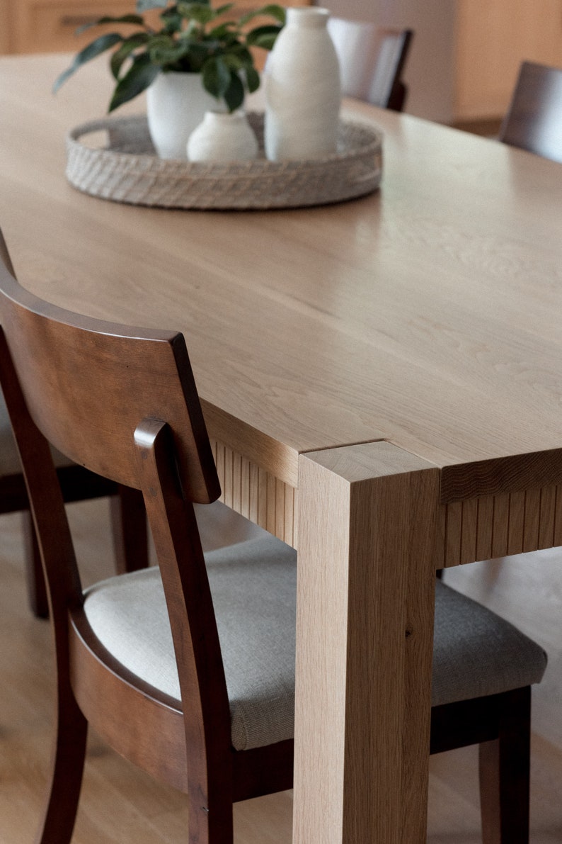 Puede incluir: Una mesa de comedor de madera con un acabado en madera clara y una bandeja tejida con jarrones de cer&aacute;mica blanca en la parte superior. La mesa tiene una forma rectangular y est&aacute; rodeada por cuatro sillas con marcos de madera marr&oacute;n y asientos de tela gris claro.