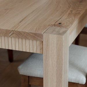 Puede incluir: Primer plano de una mesa de comedor de madera clara con un detalle de borde de listones. La mesa tiene una pata cuadrada y se muestra con una silla.