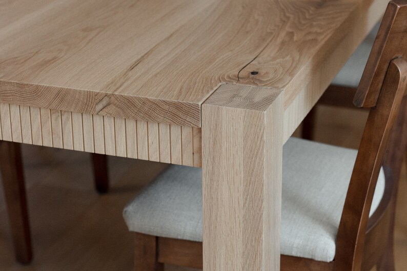 Puede incluir: Primer plano de una mesa de comedor de madera clara con un detalle de borde de listones. La mesa tiene una pata cuadrada y se muestra con una silla.