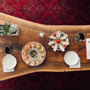 May include: A wooden table with a live edge, set for a dinner party. There are two plates of appetisers, a plate of bread with cheese and fruit, a glass of wine, a candle, and a cookbook titled "Big Macs & Burgundy".