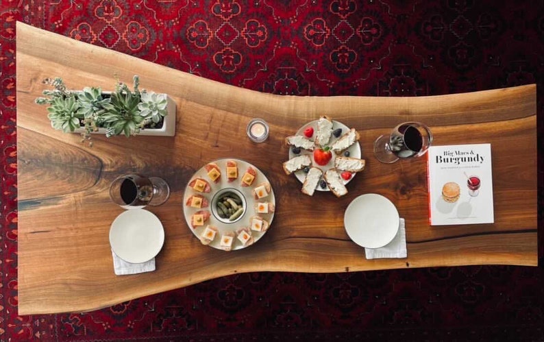 May include: A wooden table with a live edge, set for a dinner party. There are two plates of appetisers, a plate of bread with cheese and fruit, a glass of wine, a candle, and a cookbook titled "Big Macs & Burgundy".