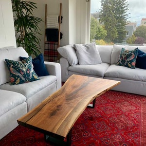 May include: A large, live edge coffee table made of wood with a natural, brown finish. The table has a unique, organic shape and is supported by black metal legs. The table is sitting on a red rug in a living room setting.
