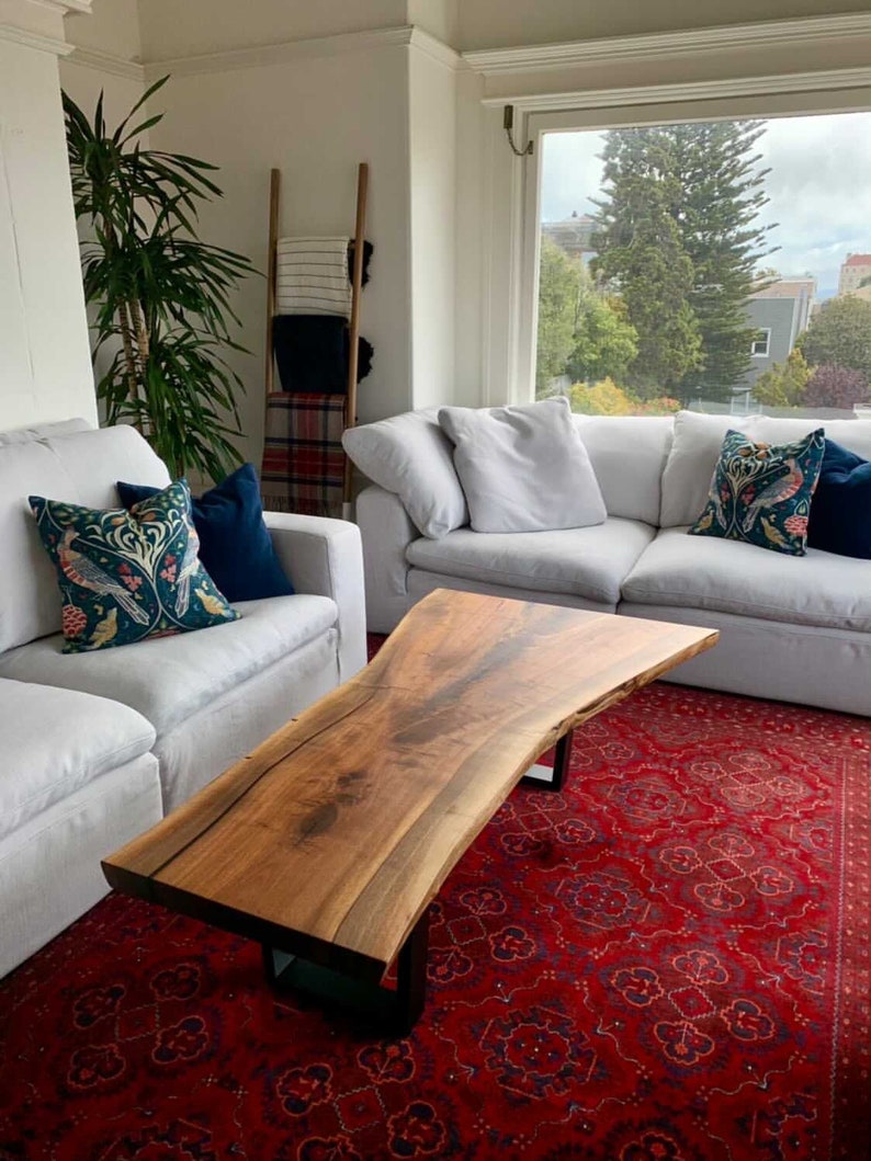 May include: A large, live edge coffee table made of wood with a natural, brown finish. The table has a unique, organic shape and is supported by black metal legs. The table is sitting on a red rug in a living room setting.