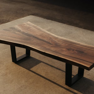 May include: A long, wooden coffee table with a live edge top and black metal legs. The table has a natural wood finish and a unique, organic shape.