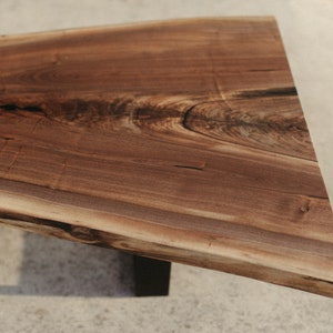 May include: A large, live edge wooden table top with a natural brown finish. The table top has a unique, irregular shape and is supported by a black metal base.