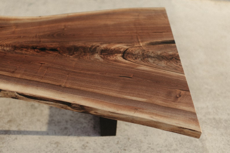 May include: A large, live edge wooden table top with a natural brown finish. The table top has a unique, irregular shape and is supported by a black metal base.