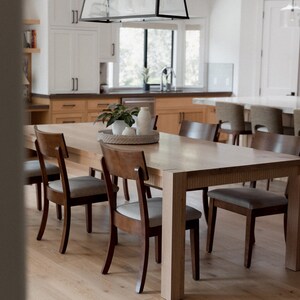 Puede incluir: Una mesa de comedor de madera clara con seis sillas. La mesa tiene un acabado de madera natural y las sillas tienen un acabado de madera oscura. La mesa est&aacute; puesta con un centro de mesa blanco y un jarr&oacute;n blanco con plantas verdes.