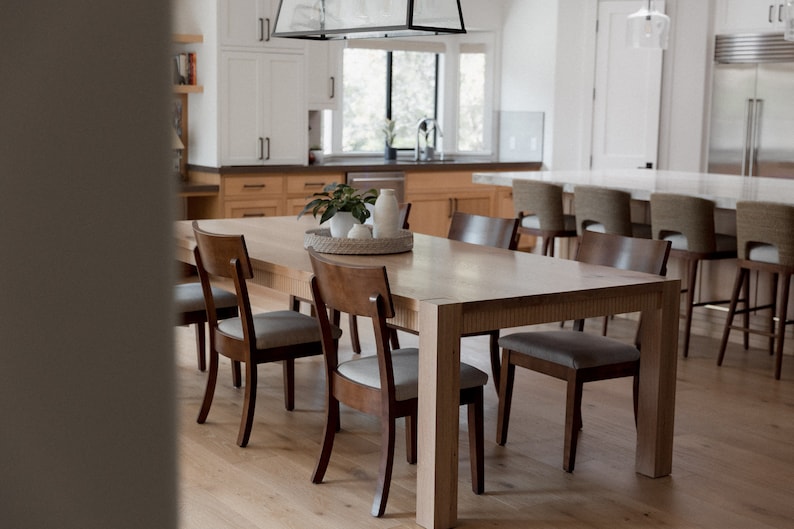 Puede incluir: Una mesa de comedor de madera clara con seis sillas. La mesa tiene un acabado de madera natural y las sillas tienen un acabado de madera oscura. La mesa est&aacute; puesta con un centro de mesa blanco y un jarr&oacute;n blanco con plantas verdes.