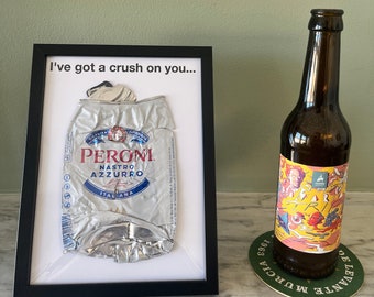May include: A framed print with the text "I've got a crush on you..." and a crushed silver can of Peroni Nastro Azzurro beer. The can is Italian and has a blue and white label. The print is on a white background with a black frame.