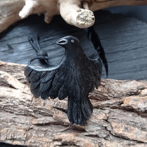 Puede incluir: Un brazalete de cuervo negro con alas detalladas y un ojo blanco. El brazalete está hecho de arcilla polimérica y se muestra sobre un trozo de madera flotante.