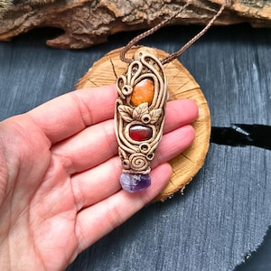 May include: A handcrafted pendant necklace featuring a beige, carved design with an orange stone, a red stone, and a purple crystal. The pendant is suspended from a brown cord, showcasing a unique, earthy aesthetic. The necklace is displayed on a wooden surface.